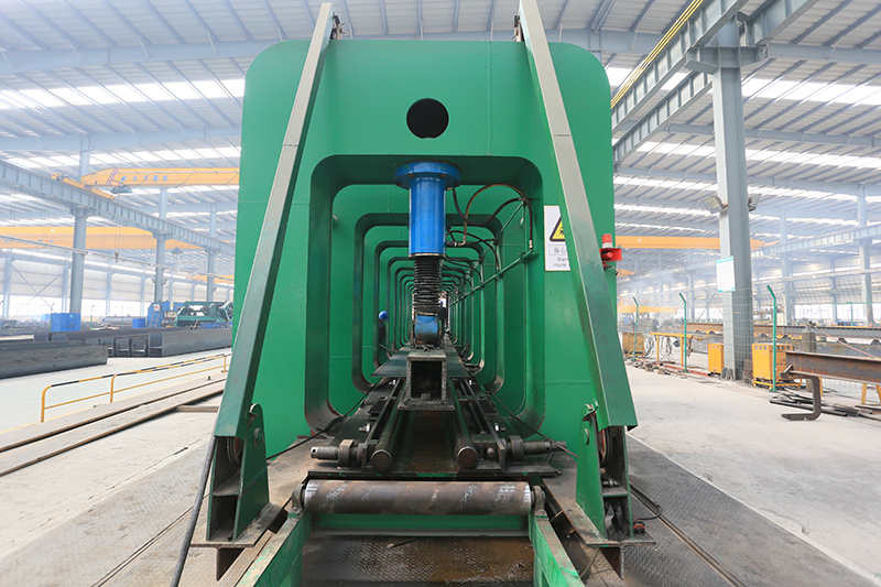 Large green industrial machine with blue hydraulic cylinder inside a warehouse, empty of people